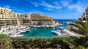 A marina with boats docked, surrounded by multi-story buildings with balconies, under a clear blue sky. - Duplex Luxury Apartment in Portomaso With Pool (St. Julian's)