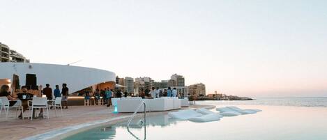 An infinity pool with lounge chairs and a seating area, overlooking the sea with buildings in the background.