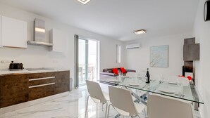 A modern kitchen with a built-in oven, a dining area with a glass table and chairs, and a living area with a sofa and a painting. - Cosy Apartment Steps to St George's Bay (Swieqi)
