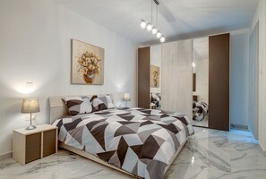A modern bedroom with a geometric-patterned bedspread, a bedside lamp, a mirror, and a painting on the wall. - Cosy Apartment Steps to St George's Bay (Swieqi)
