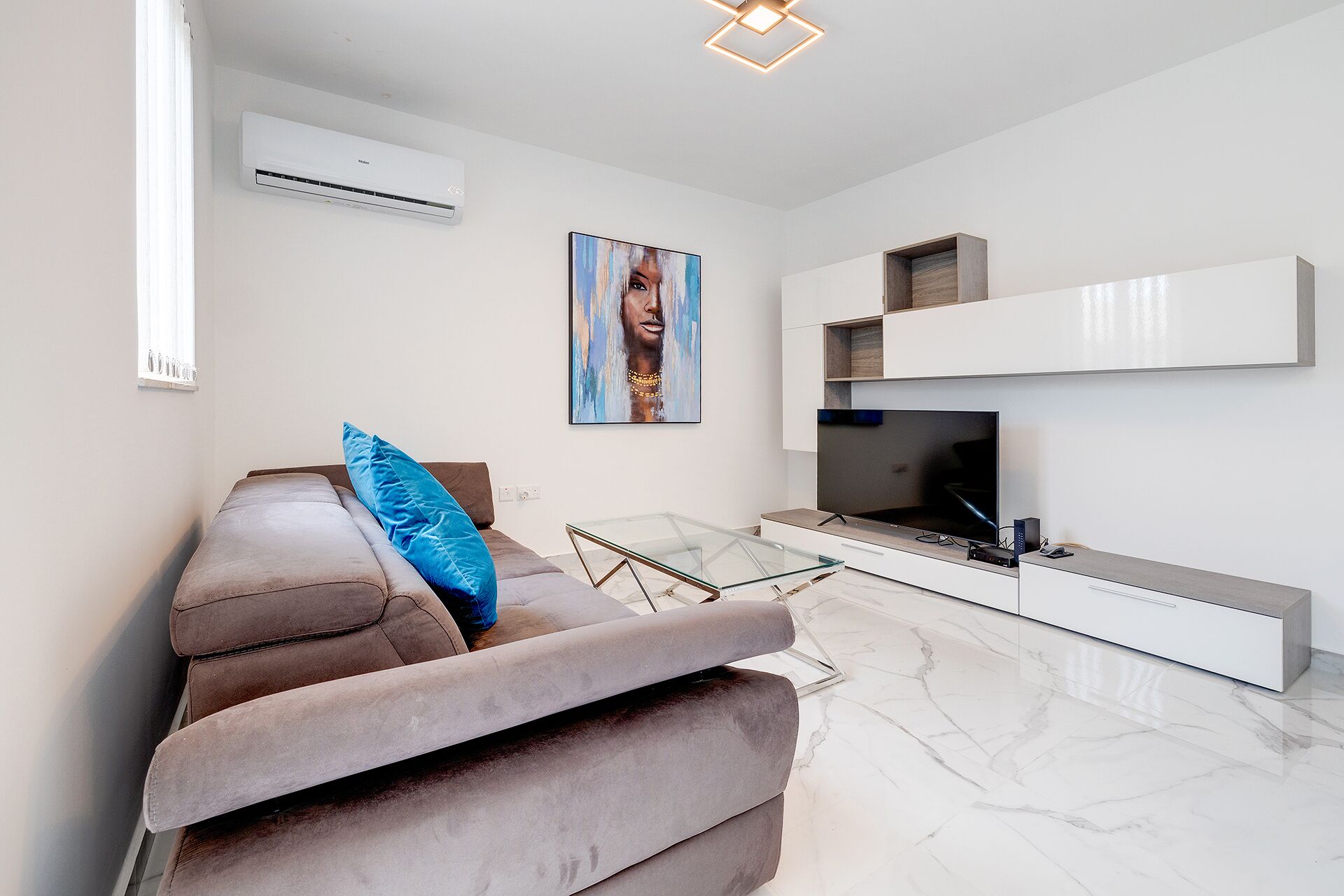 A modern living room with a sofa, a glass coffee table, a flat-screen TV, a mounted shelf, and an air conditioning unit.