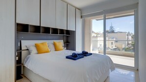 A modern bedroom with a large bed, white bedding, and yellow pillows. There is a window with a city view. - Cosy 1BR Apartment in Central St Julian's (Swieqi)