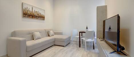 A modern living room with a sectional sofa, a dining table with chairs, a flat-screen TV, and a cityscape painting on the wall.