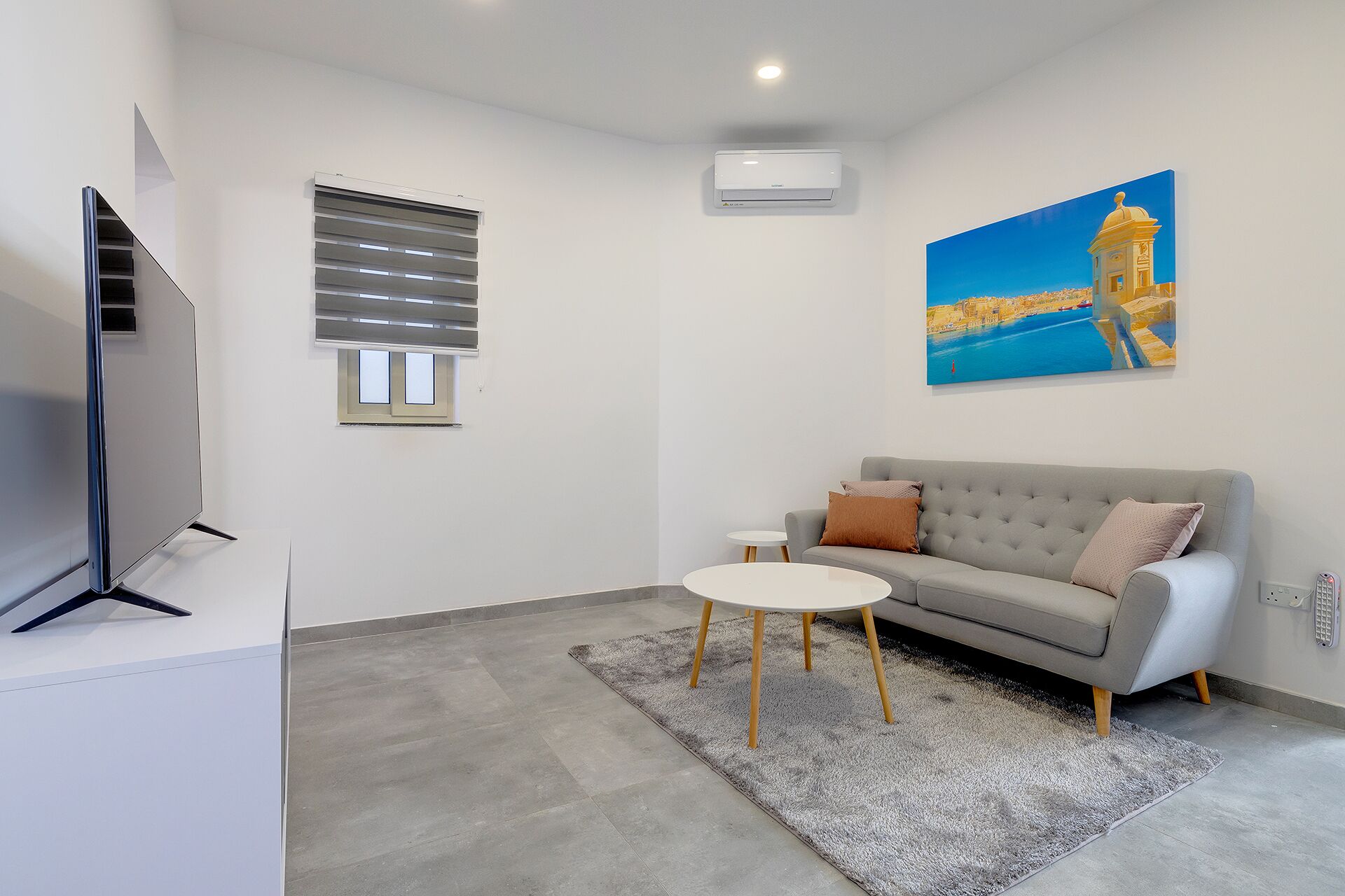 A modern living room with a grey sofa, a small white table, a flat-screen TV, a painting of a coastal scene, and a window with blinds.