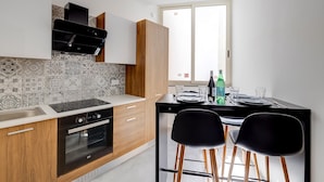 Cozinha moderna com forno embutido, fogão e pia. Existe uma área de jantar com duas cadeiras e uma mesa posta para dois.