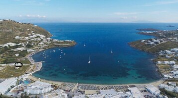 Vista aérea de una zona costera con aguas azules cristalinas, barcos y una playa con tumbonas y sombrillas.