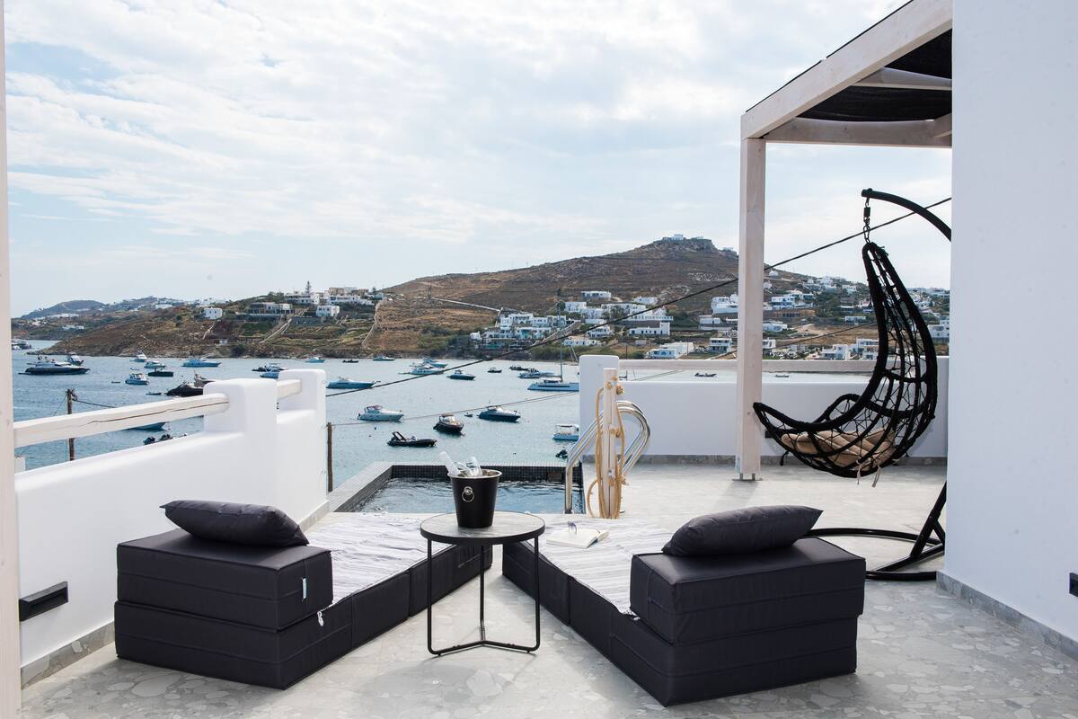 A balcony with two black sofas, a small table, and a hanging chair overlooking a marina with boats and a hilly town in the background.