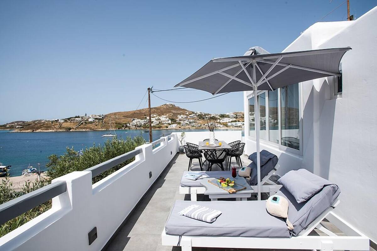 A balcony with a white railing, a table with chairs, a sun umbrella, and lounge chairs with cushions.