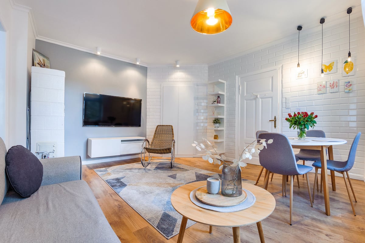 A modern living room with a sofa, a dining table with chairs, a TV mounted on the wall, and a decorative vase on the coffee table.