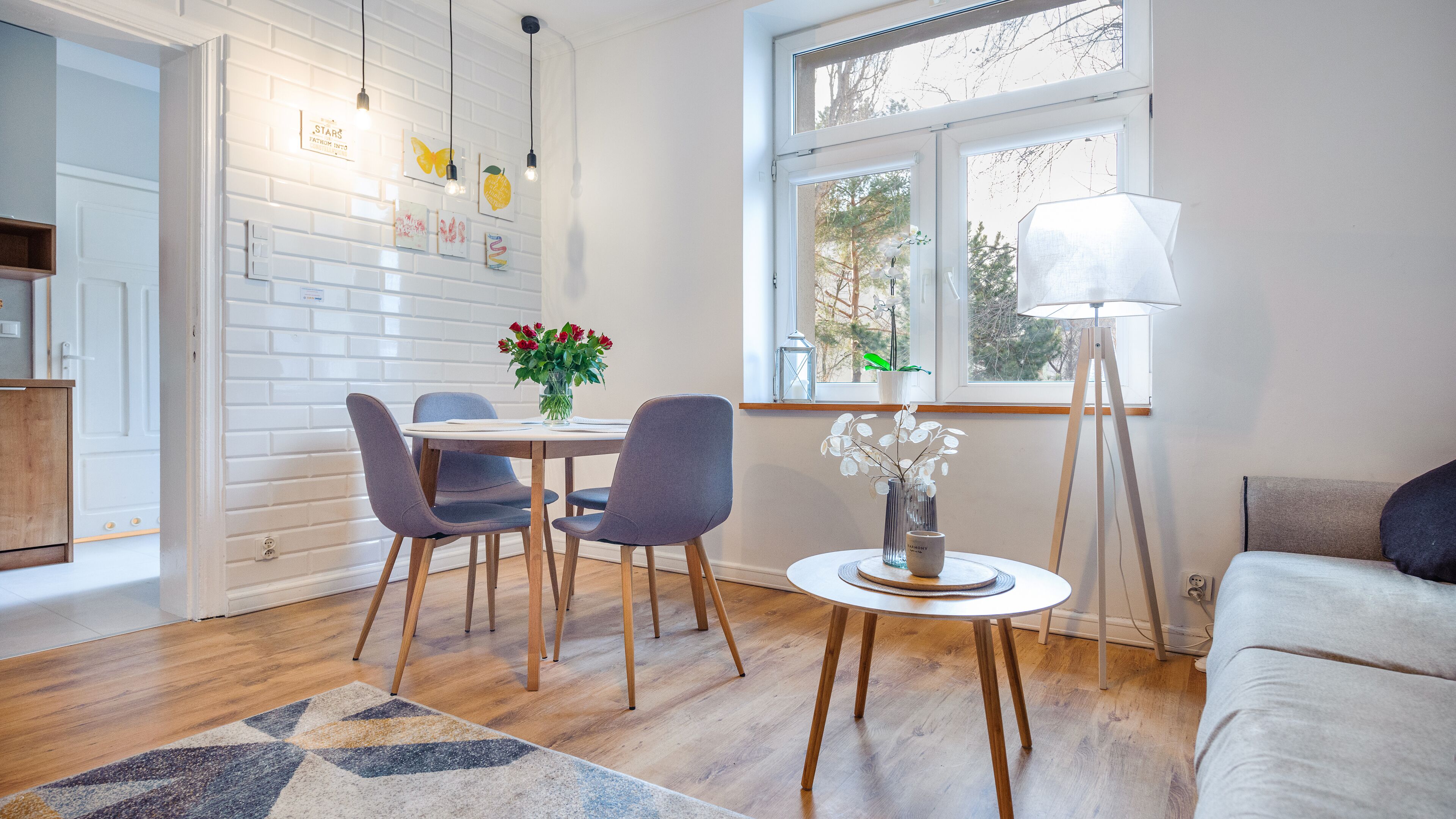 Un salon moderne avec un coin repas, caractérisé par un mur en briques blanches, un plancher en bois et une grande fenêtre donnant sur la verdure.