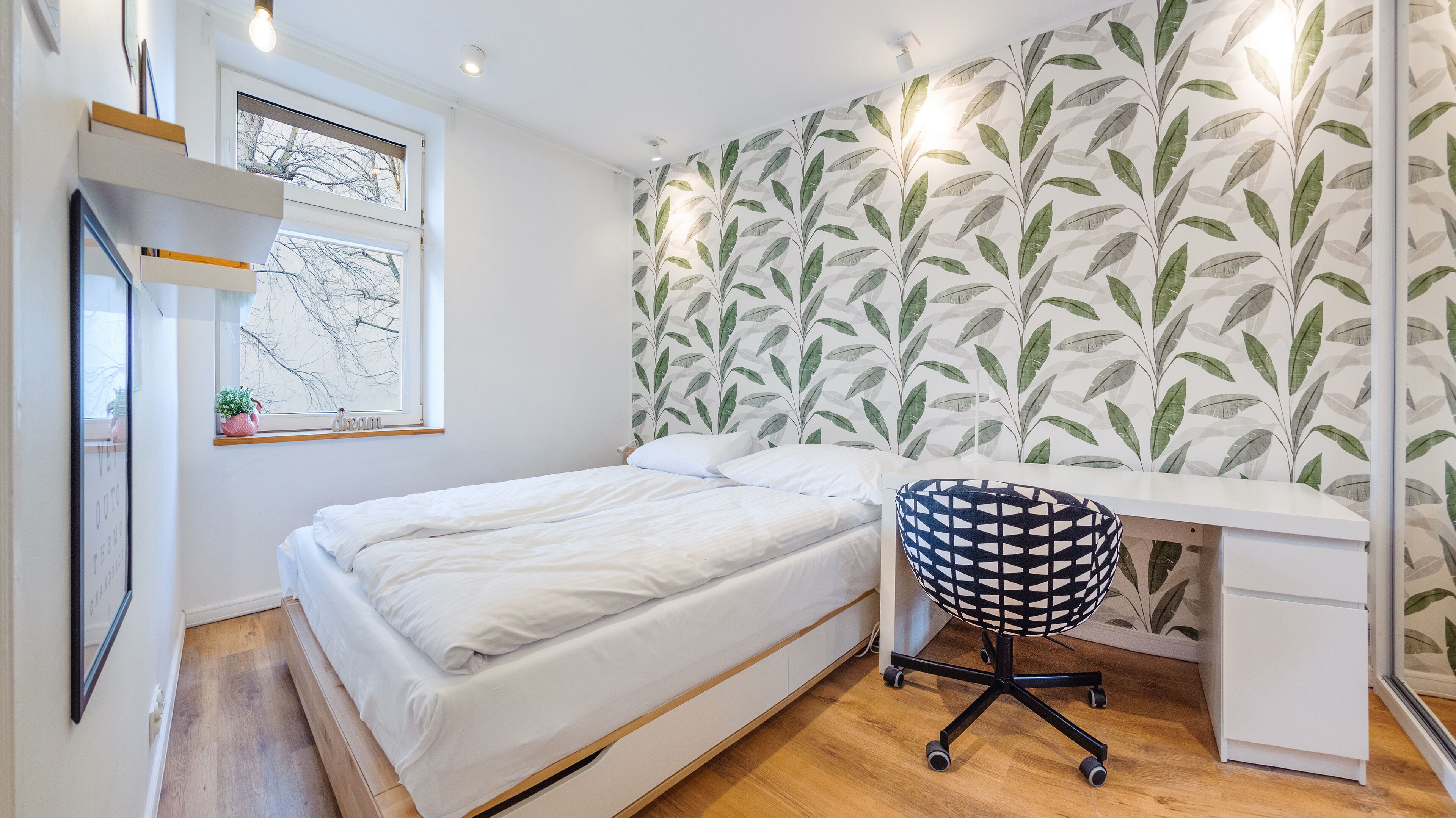 A bedroom with a bed, a desk, and a chair. There is a window with a view of bare trees, a wall with a leaf pattern, and a shelf with items.