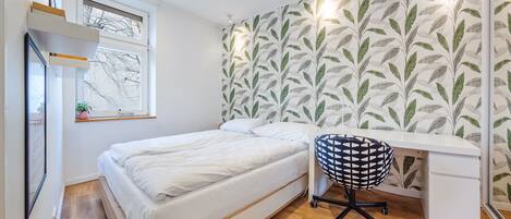 A bedroom with a bed, a desk, and a chair. There is a window with a view of bare trees, a wall with a leaf pattern, and a shelf with items.