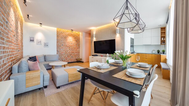 Een moderne woonkamer met een bakstenen muur, een bank, een eettafel gedekt met borden en glazen, en een keukenhoek met kasten en een televisie.