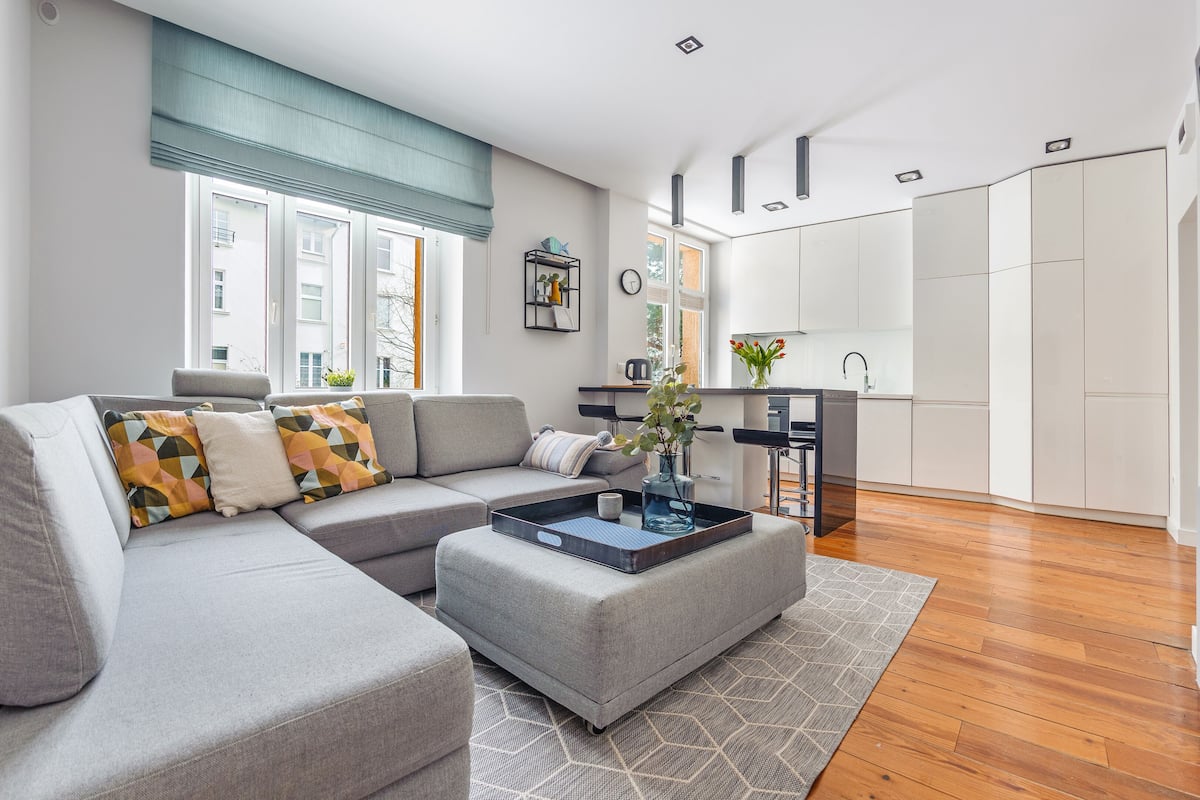 A modern living room with a grey sectional sofa, a coffee table, and a kitchen area with white cabinets and a black countertop.