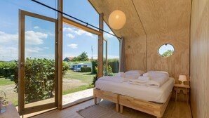 Een slaapkamer met een houten bed, een ronde spiegel en uitzicht op een tuin en geparkeerde auto door grote glazen deuren.