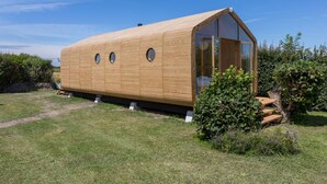 Een houten, langgerekt gebouw met een puntig dak en grote ramen, gelegen in een grasrijk gebied met een heldere lucht.