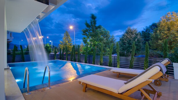 A pool area with a waterfall feature, lounge chairs, and a view of trees and a fence. - Selin Luxury Residences (Ioannina)