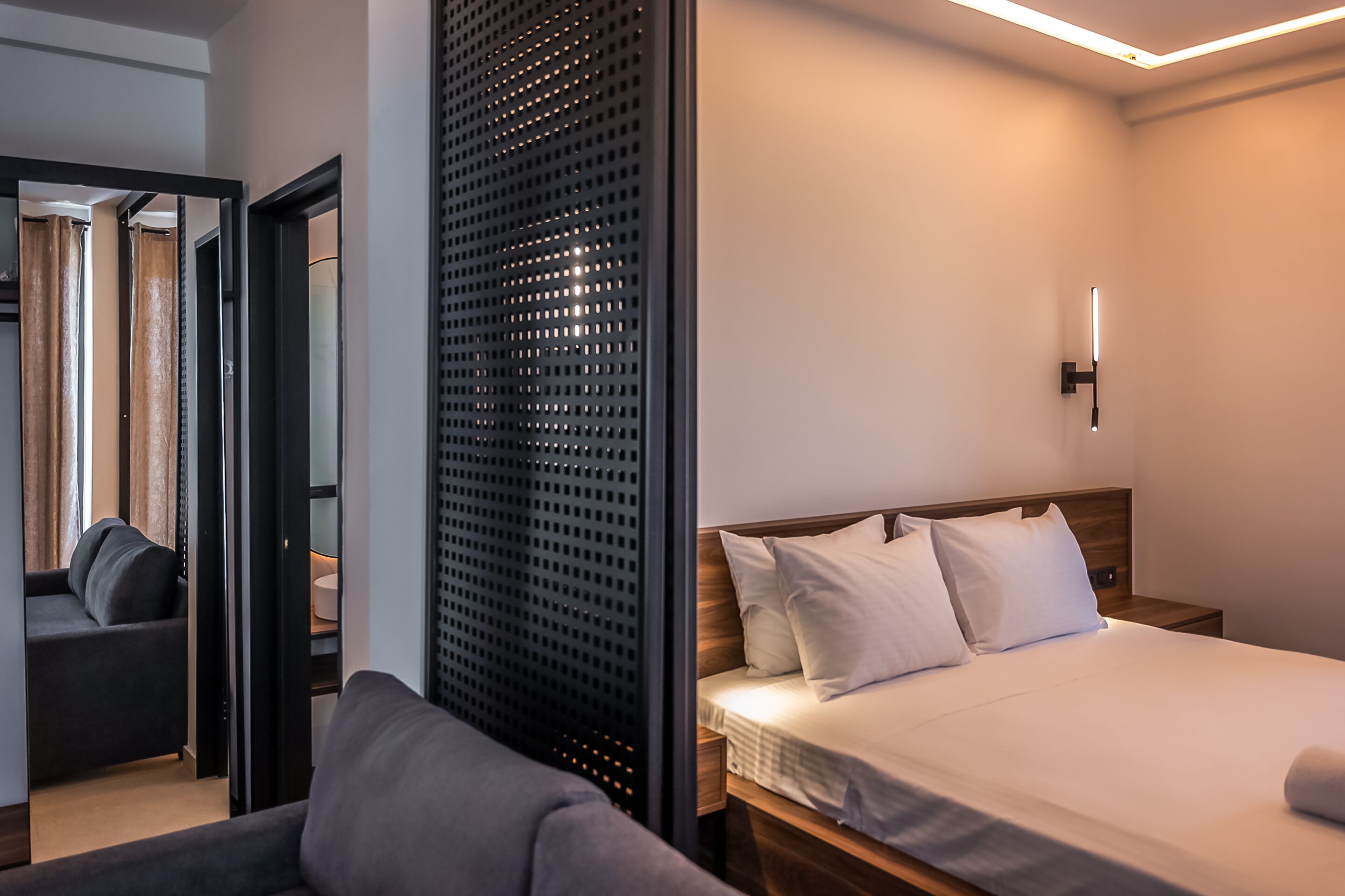 A hotel room with a bed, a sofa, a wooden headboard, a wall-mounted light, and a door leading to another room.