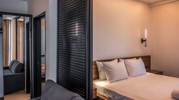 Una habitación de hotel con una cama, un sofá, cabecera de madera, una lámpara de pared y una puerta que da a otra habitación.