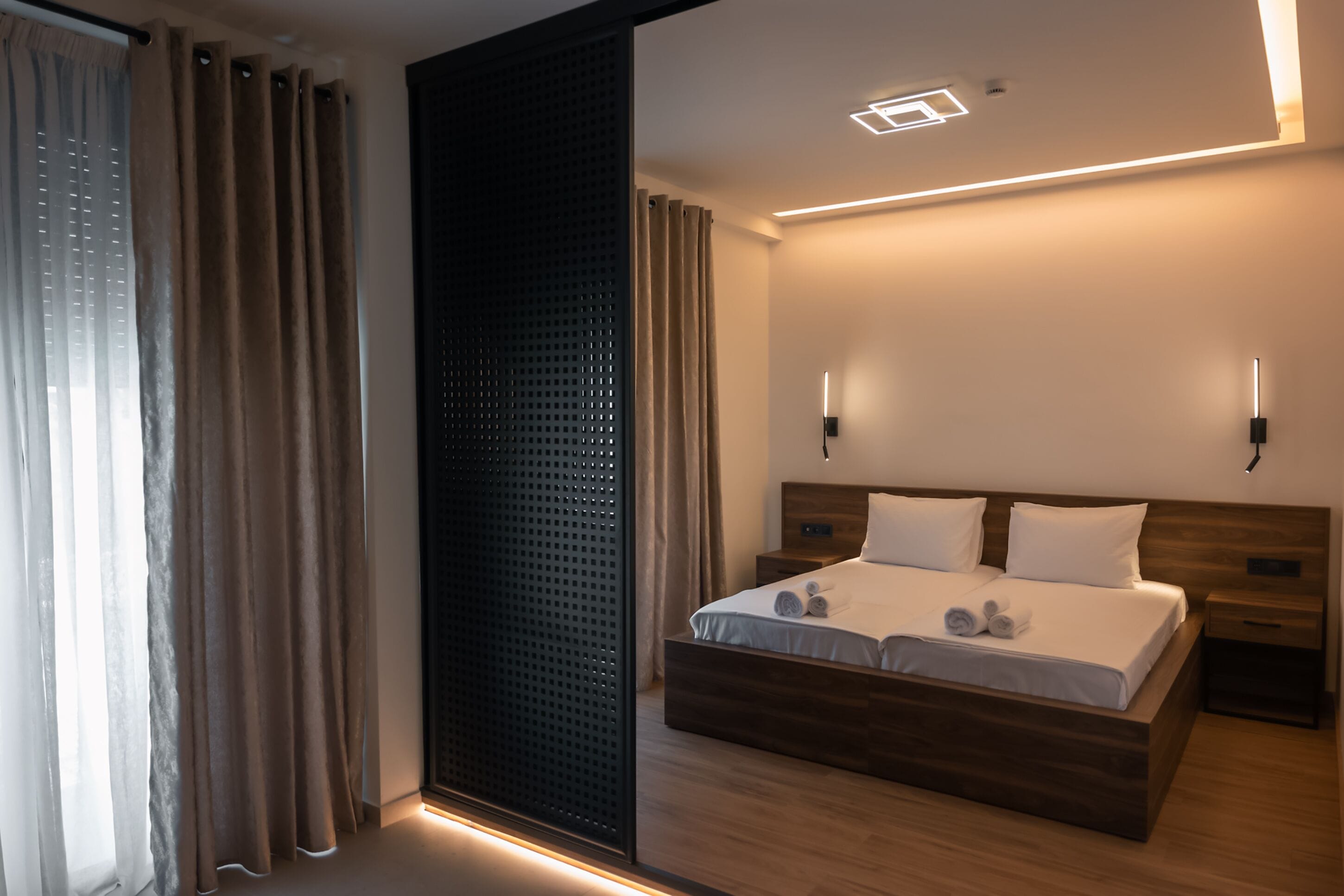 Une chambre d’hôtel moderne avec un lit en bois, deux tables de chevet, un grand miroir et un luminaire fixé au mur.
