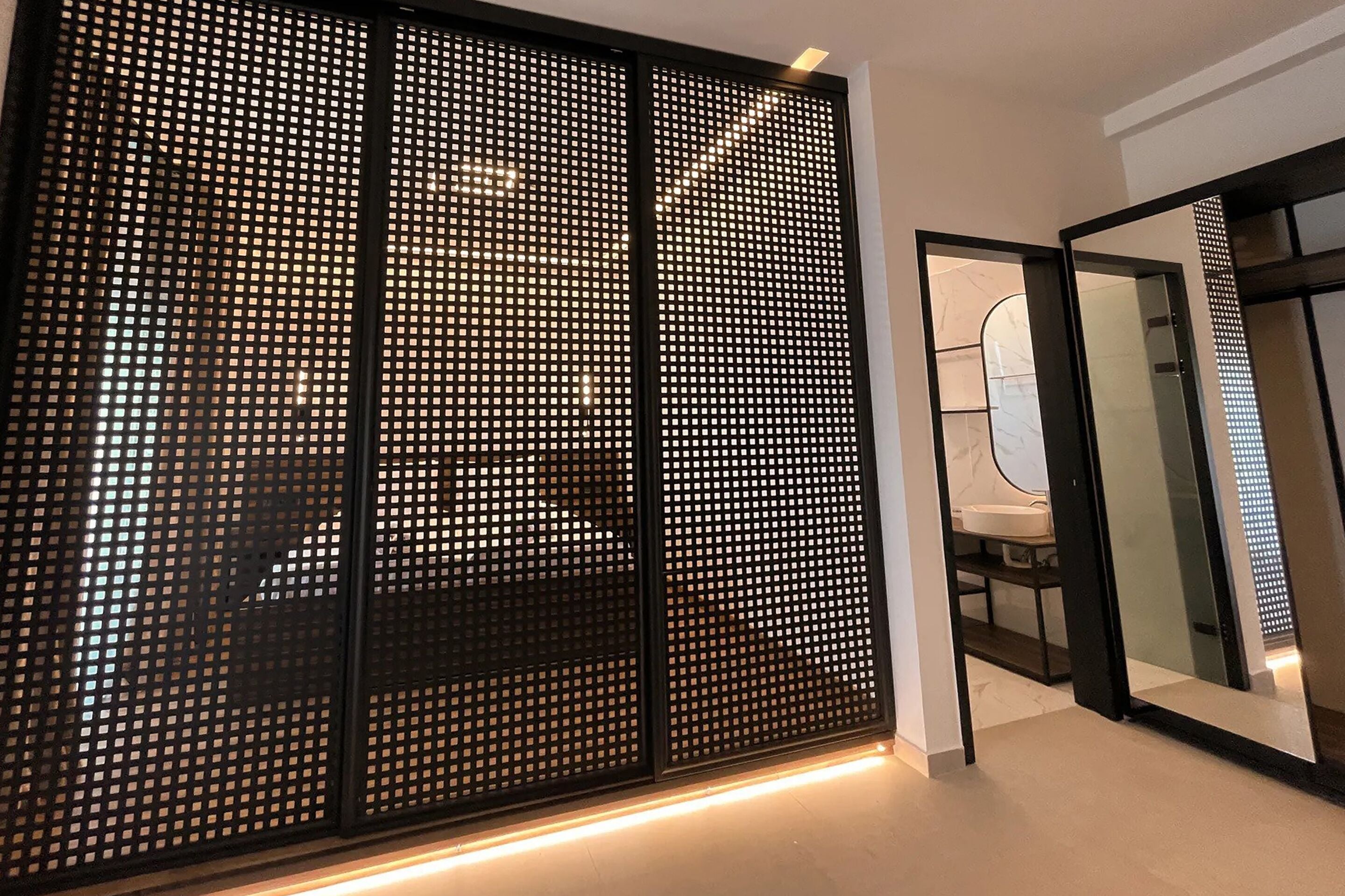 A modern room with a sliding black perforated partition, a glass door leading to a bathroom, and a white sink.