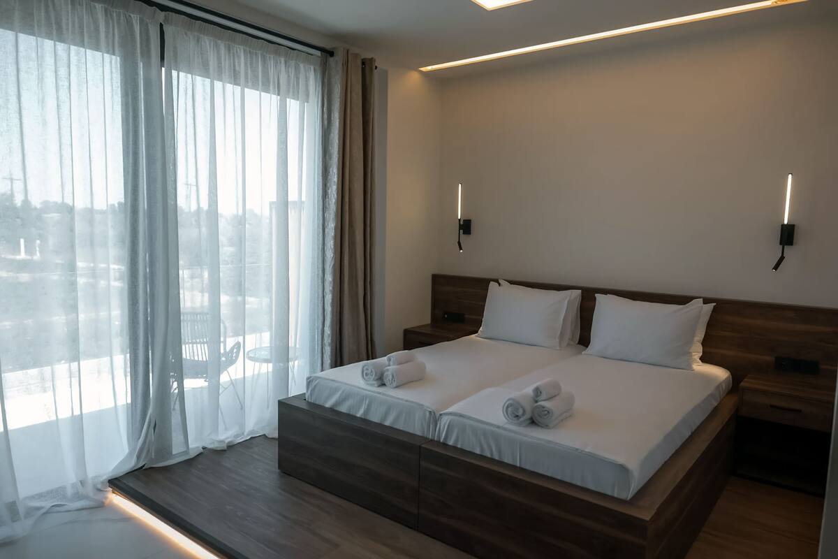 Una cama doble con sábanas blancas, dos mesitas de noche, un cabecero de madera y vistas a través de cortinas de gasa.