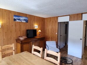 A room with a wooden wall, a television, a lamp, a dining table with chairs, and a door leading to another room.