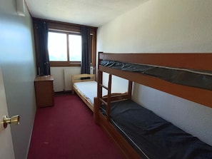 A small, compact room with a bunk bed, a single bed, a small table, and a window with curtains.