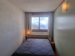 Een kleine, smalle kamer met een enkel bed, een raam met gordijnen en een nachtlampje.