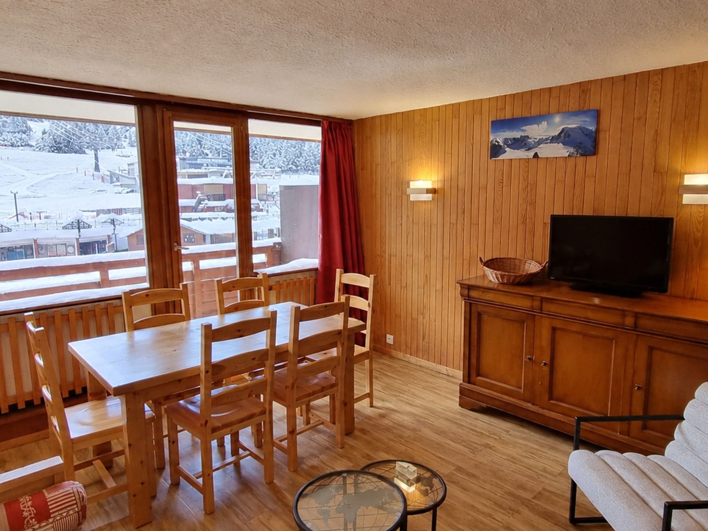 A wooden-paneled room with a dining table, chairs, a television, and a view of a snowy landscape through large windows.