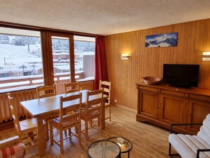 Een kamer met houten lambrisering, een eettafel, stoelen, een televisie en uitzicht op een besneeerd landschap door grote ramen.