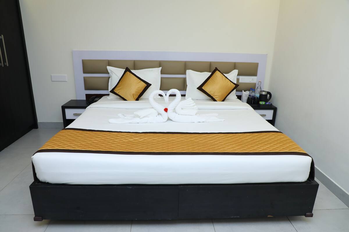 A neatly made bed with a swan towel design, a bedside table with a phone and a bottle, and a nightstand with a lamp.
