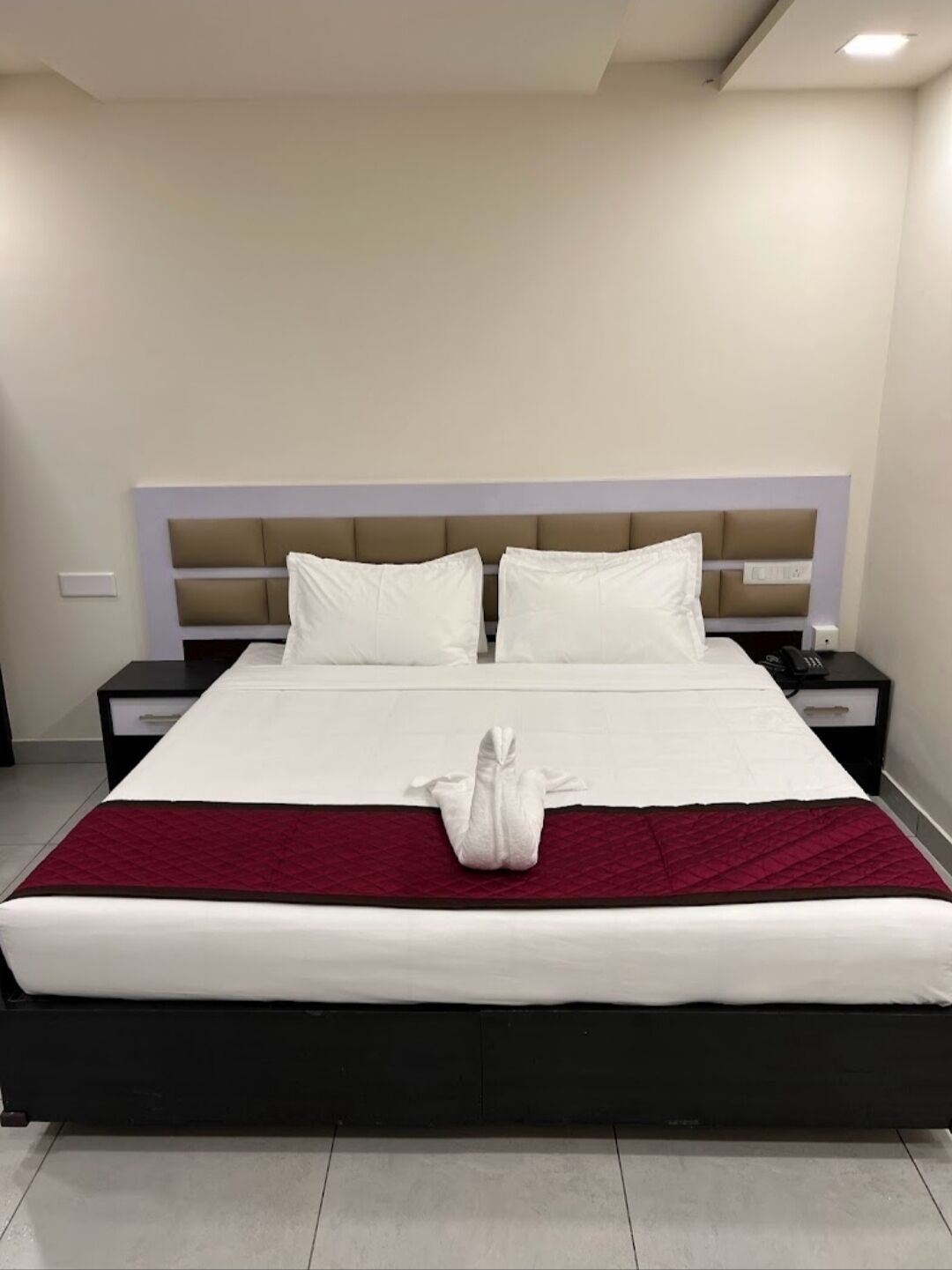 A hotel room with a large bed, white bedding, a red throw blanket, and a white towel on the bed.