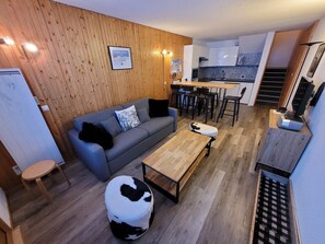 Un salon moderne avec un canapé gris, une table basse en bois et un pouf à motifs de vaches. On aperçoit à l’arrière-plan une cuisine avec des meubles blancs et un dosseret décoré.