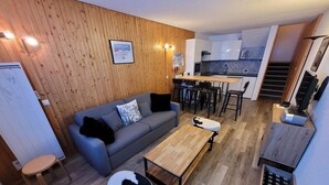 Ein modernes Wohnzimmer mit einem grauen Sofa, einem Holztisch und einem Ottomanen mit Kuhmuster. Im Hintergrund ist eine Küche mit weißen Schränken und einer gemusterten Spritzschutzwand erkennbar.