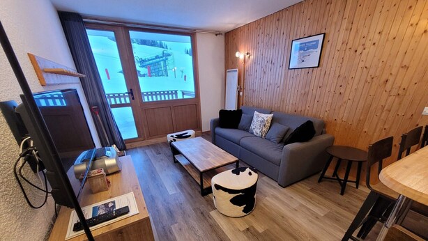 Ein gemütliches Wohnzimmer mit einer Couch, einem Couchtisch und einem kleinen runden Tisch. Eine Holzwand und ein Fenster mit Blick auf eine verschneite Landschaft.
