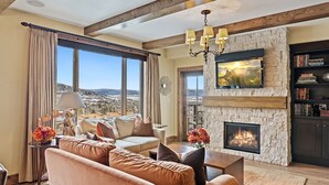 A cozy living room with a stone fireplace, a large TV mounted on the wall, and a view of the mountains through the windows.