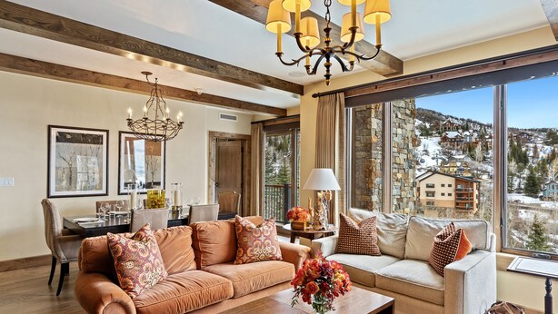 A spacious living room with a large window offering a view of a snowy landscape, a wooden ceiling, and a chandelier.
