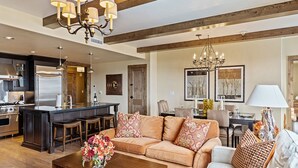 A spacious living area with a large sofa, a coffee table with flowers, a chandelier, and a kitchen in the background.