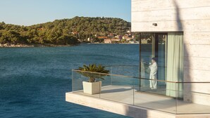 Un edificio moderno con balcón de vidrio que da a una masa de agua y una colina con casas.