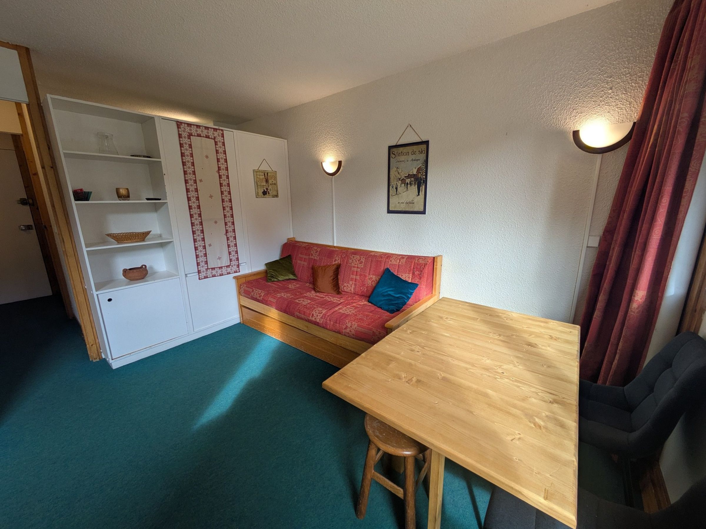 A cozy room with a wooden table, a red patterned sofa, a white cabinet with shelves, and a wall-mounted light fixture.
