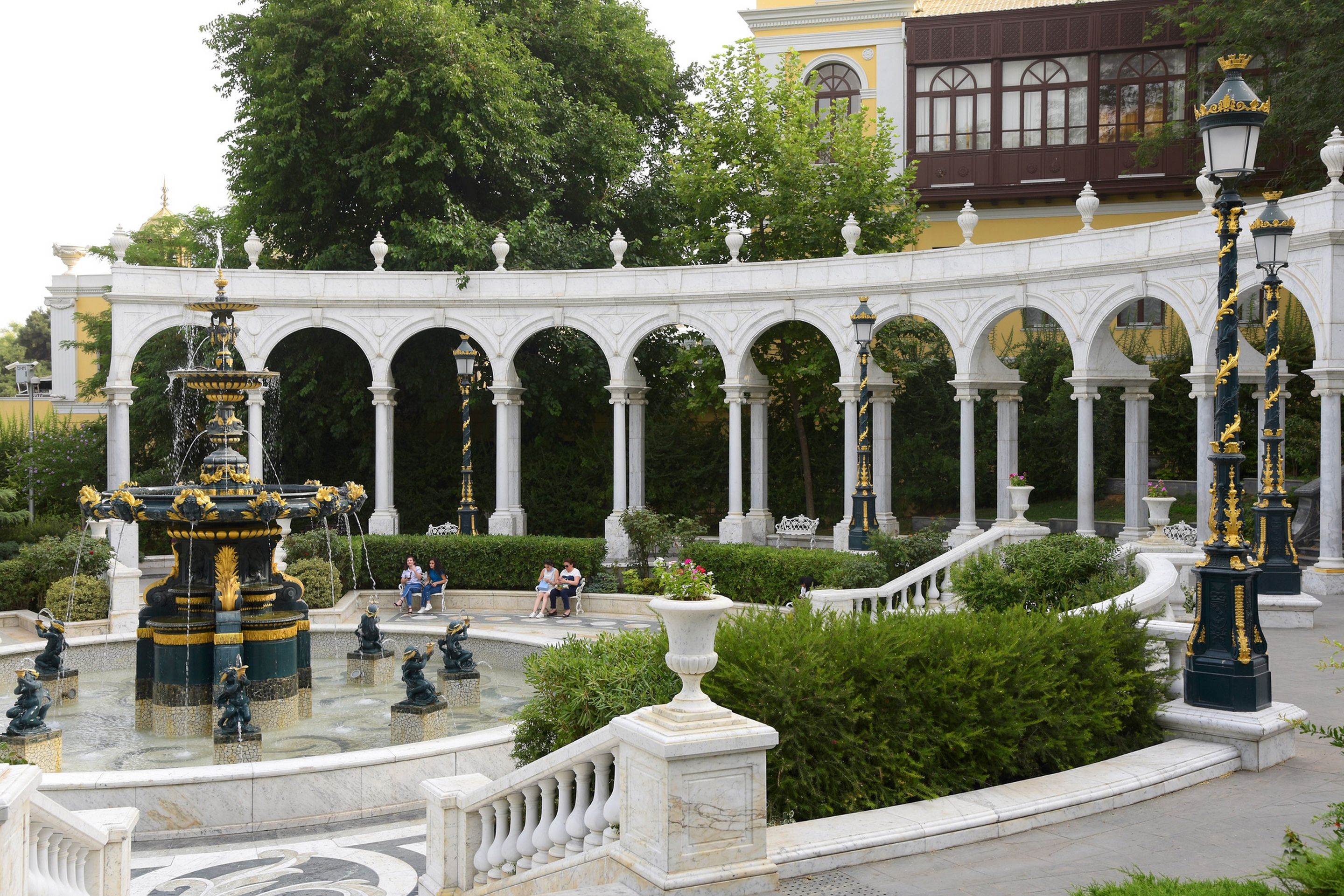 A classical garden with a fountain, white columns, and ornate lamp posts.
