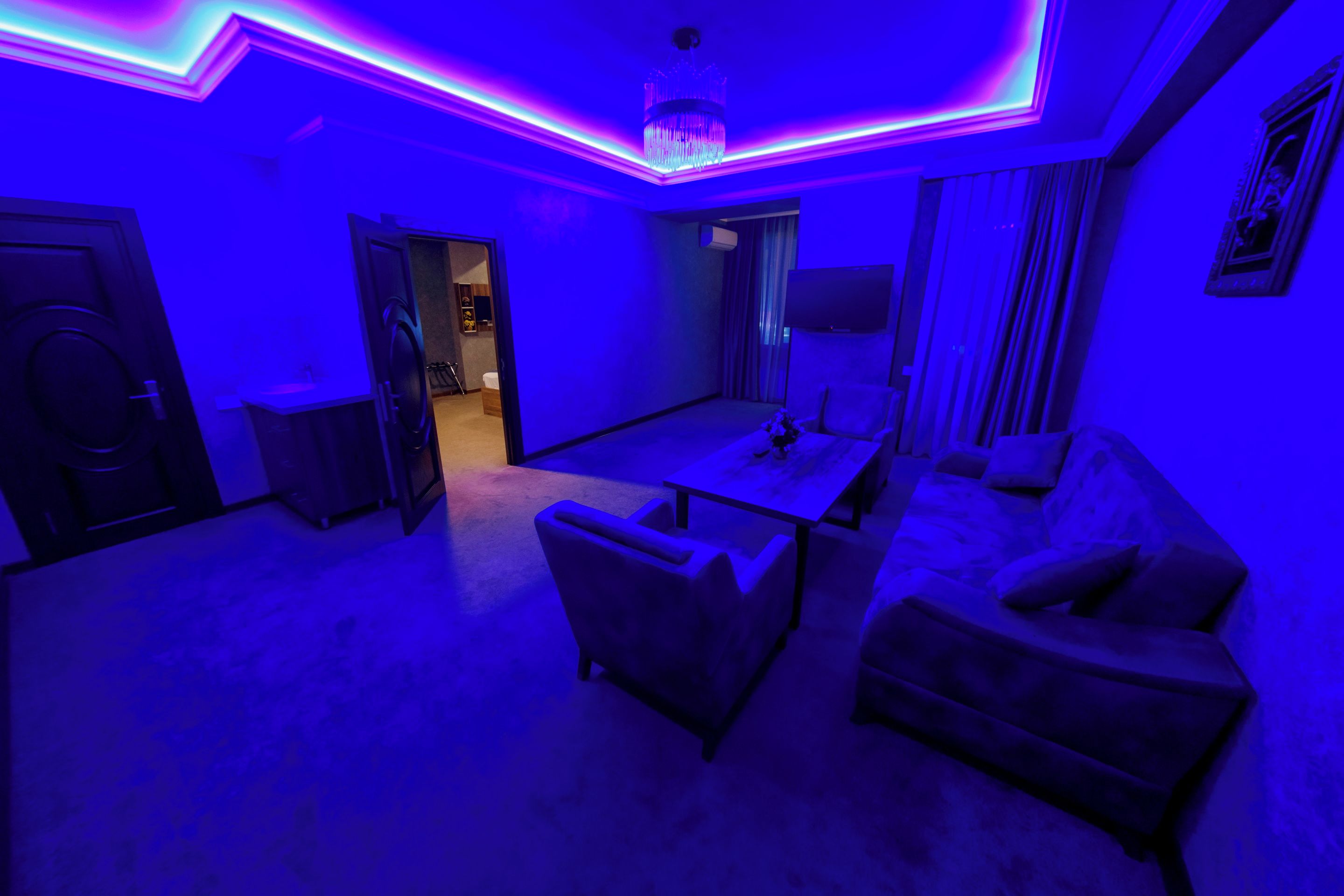 Un salon moderne avec un canapé, un fauteuil et une table basse. Un téléviseur est fixé au mur et un lustre est suspendu au plafond.