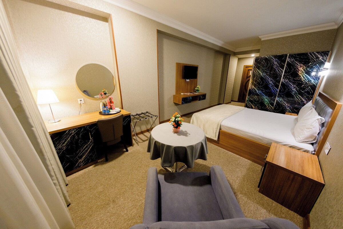 A hotel room with a bed, a desk, a chair, a small table, a mirror, and a TV.