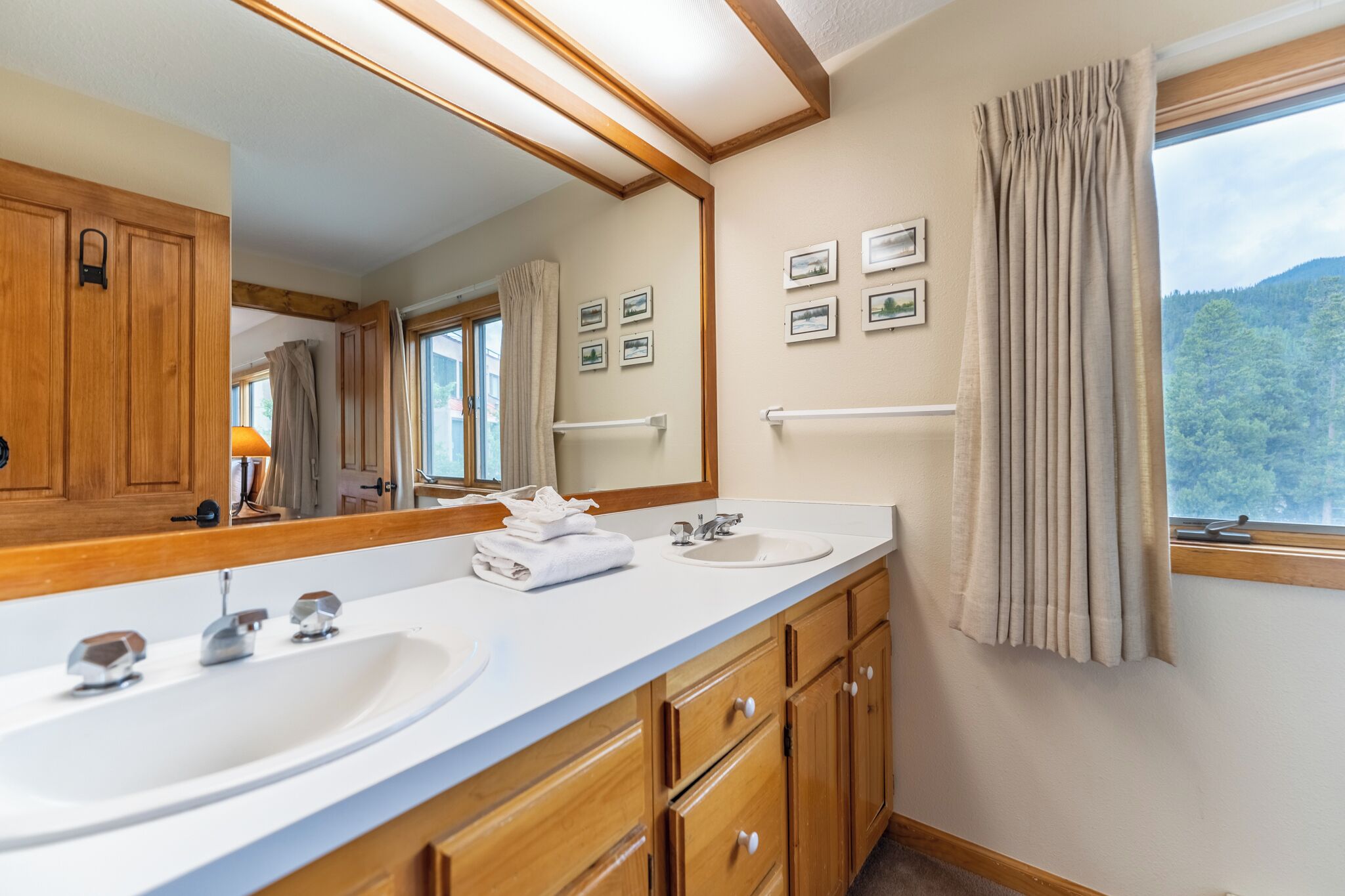 A bathroom with two sinks, wooden cabinets, a large mirror, and a window with curtains.