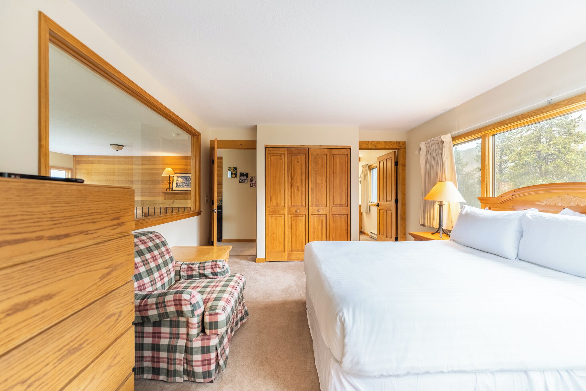 A bedroom with a large bed, a wooden wardrobe, a plaid chair, and a nightstand with a lamp.