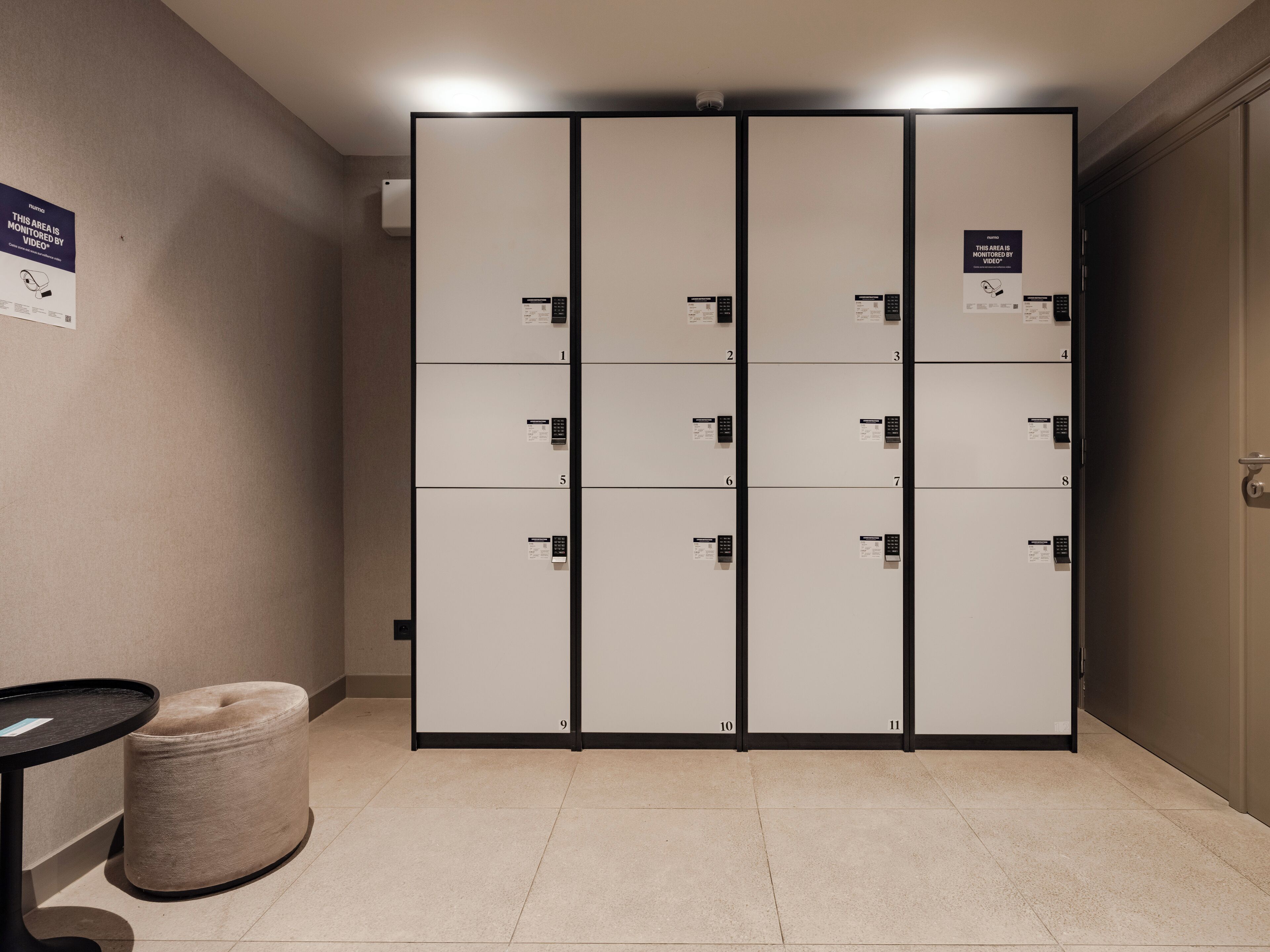A row of hotel lockers with numbered doors, a small round ottoman, and a table with a placard on the wall.