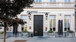 Un bâtiment avec une porte d’entrée noire, une enseigne indiquant « numastays.com » et des parasols à l’extérieur.
