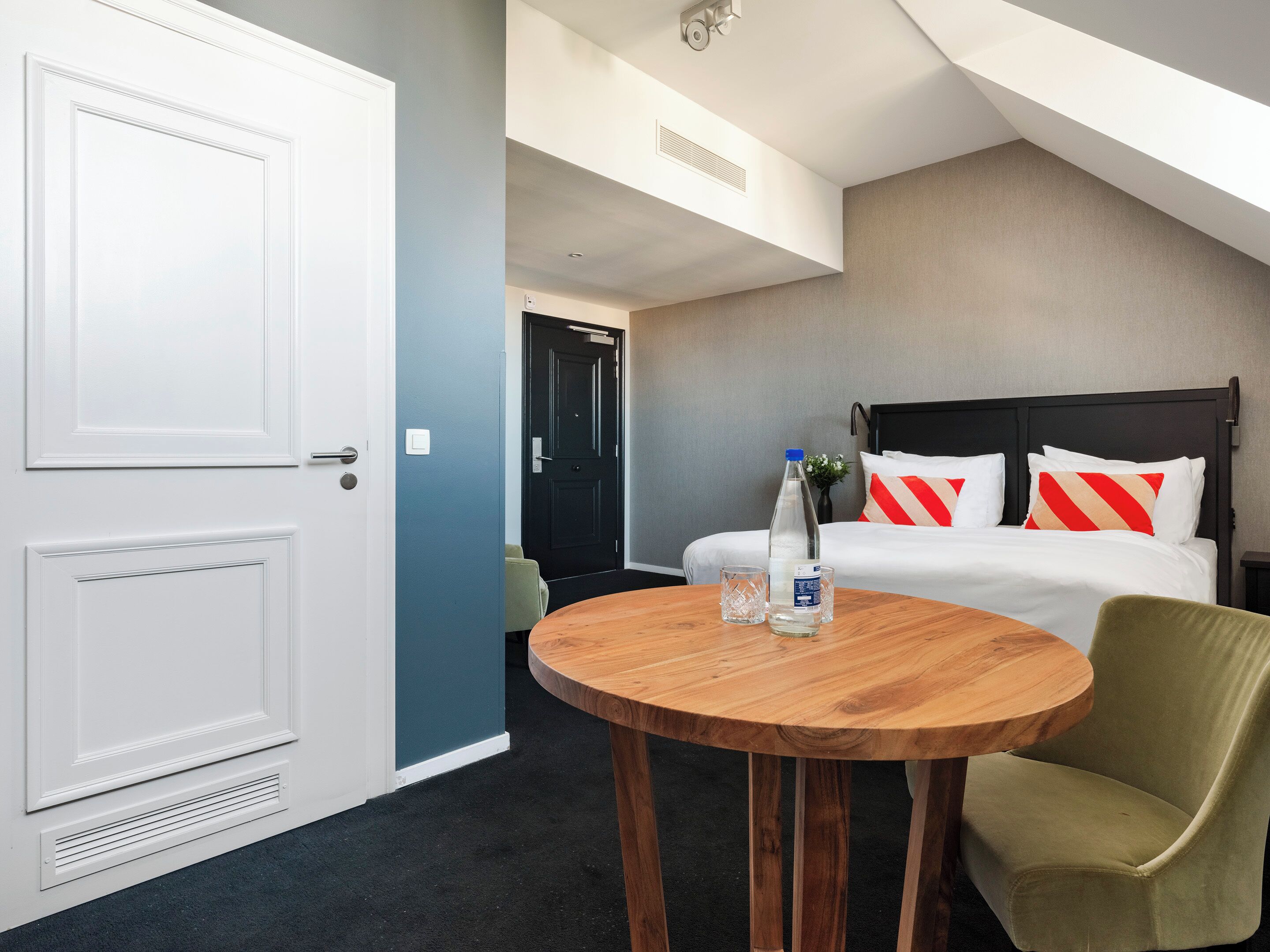 A hotel room with a bed, a wooden table, a chair, and a door.