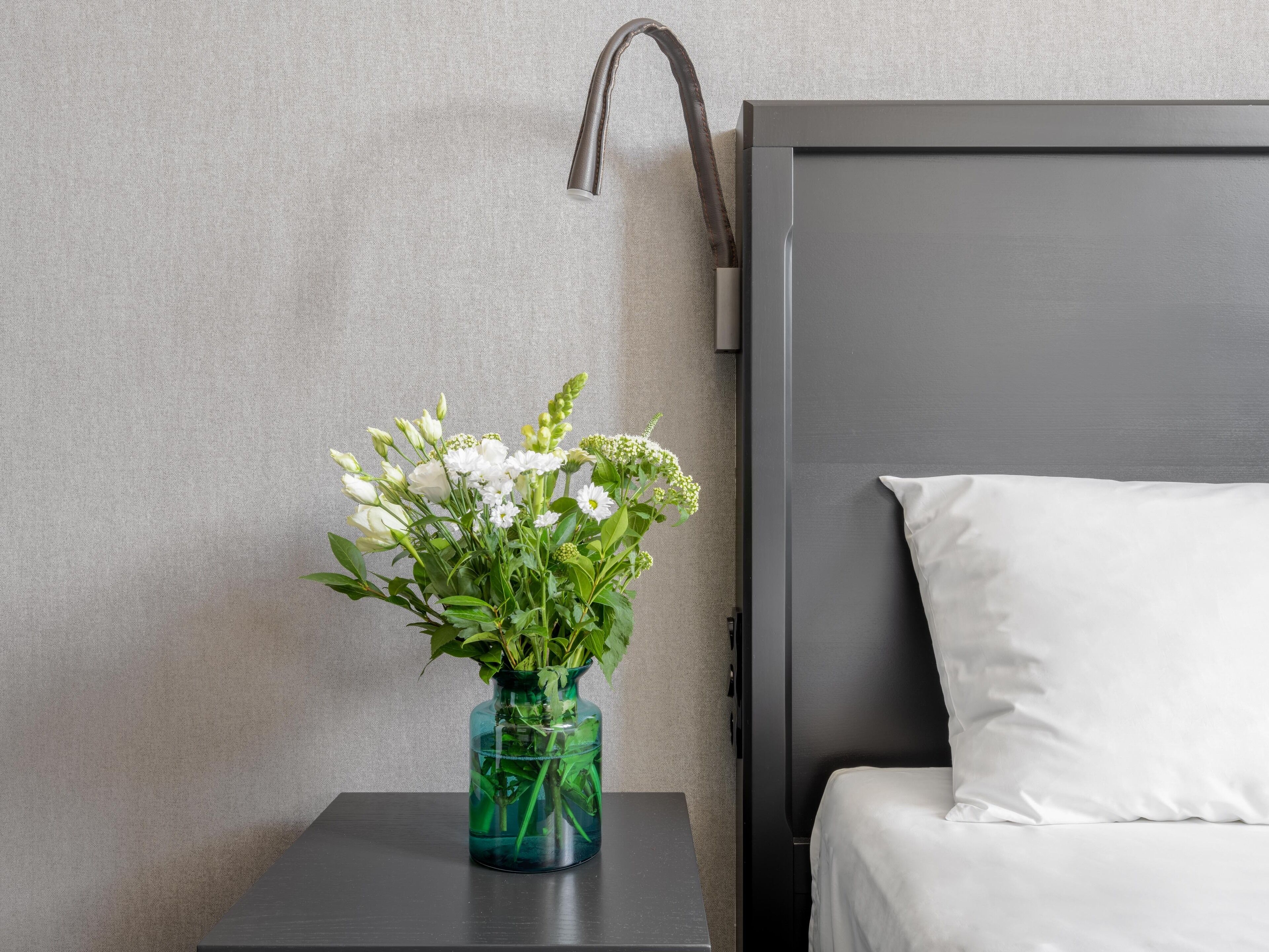 A bed with white bedding, a small table with a vase of flowers, and a wall-mounted lamp.